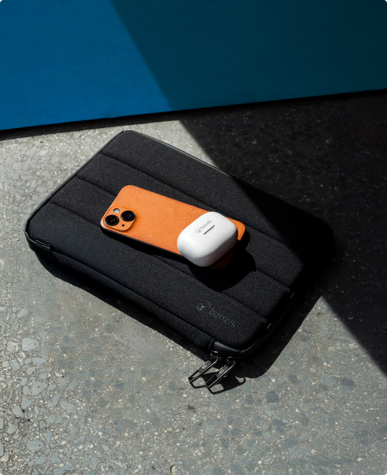 Orange iPhone and white earbuds case resting on a black Bonelk laptop sleeve in sunlight