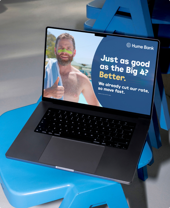 Laptop on a blue stool displaying a Hume Bank campaign ad reading “Just as good as the Big 4? Better.”