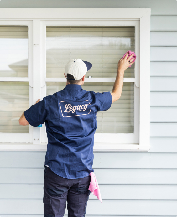 Legacy crew member in a branded uniform cleaning the exterior window of a white weatherboard home