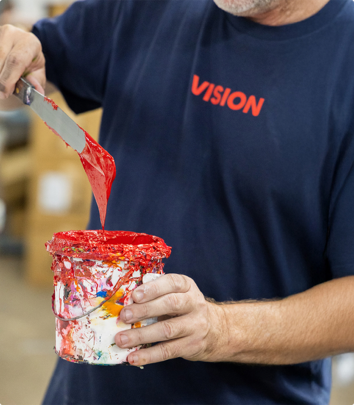Printer in a navy VISION tee scooping bright red ink from a paint tin